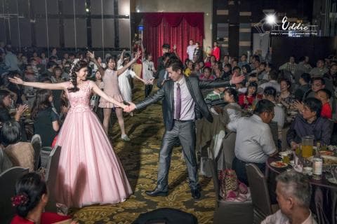 這張照片是一對新人正在跳舞，新娘穿著粉紅色的禮服，新郎穿著深灰色的西裝。他們在婚宴現場，背景有很多賓客在觀禮。