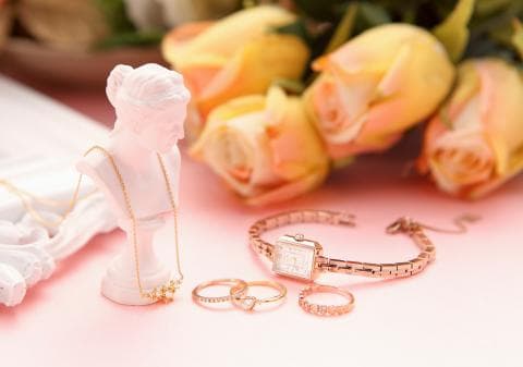 這張「珠寶和玫瑰」的照片是一個粉紅色的背景，上面放著一條金項鍊、一隻金手錶、三個金戒指和幾朵玫瑰。