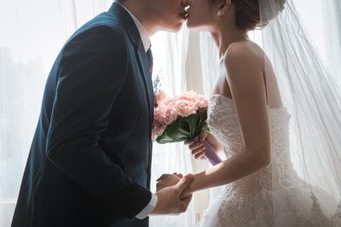 這張照片是 一對新人正在擁吻，新郎穿著黑色西裝，新娘穿著白色婚紗，他們手裡拿著一束粉紅色的花。