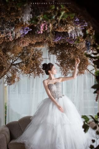 這張婚紗照是一名穿著白色蓬蓬裙婚紗的新娘，背景是枯萎的植物裝飾，她優雅地站在佈置好的攝影棚裡。