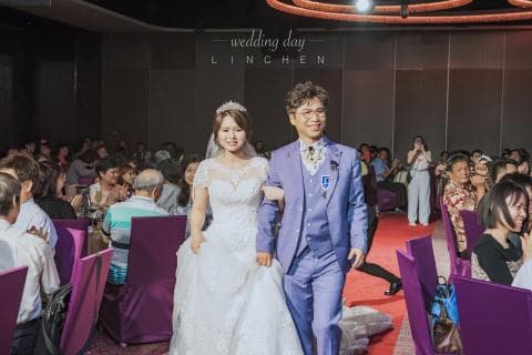 這張婚禮的照片是新人和親朋好友在婚宴現場，新郎穿著藍色西裝，新娘穿著白色婚紗，背景有紫色桌椅和金色的裝飾。