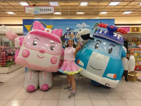 這張「卡通人物」的照片是一名女孩站在兩隻大型充氣玩偶中間，左邊是粉紅色的救護車，右邊是藍色的警車。