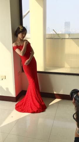 這張照片是一個身穿紅色禮服的年輕女子，她正站在窗戶前，背景是城市風景。她看起來很優雅，笑容甜美，紅色的禮服襯托出她白皙的肌膚。