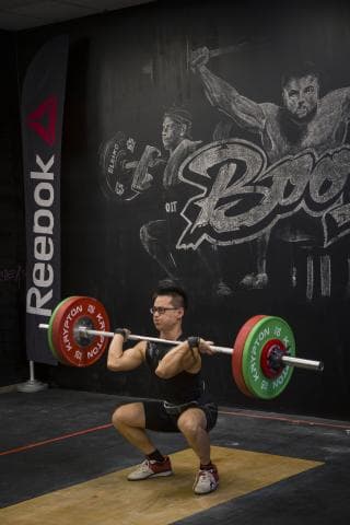 這張照片是一個年輕男子正在舉重。他正舉起一個槓鈴，上面有兩個綠色和紅色配色的重量盤。男子正在努力地舉起槓鈴，他的肌肉緊繃，臉上露出專注的表情。