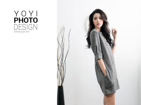 這張「時尚攝影」的照片是一位穿著灰色連身裙的年輕女性，她站在一個白色背景前，背景中有一盆裝飾性的植物。