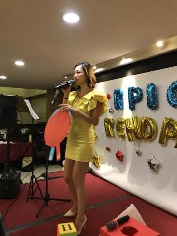 這張「一位女士在舞臺上唱歌」的照片是一名穿著黃色洋裝的女性，她拿著麥克風在唱歌，背景有一面寫著「RPIC」的牆。