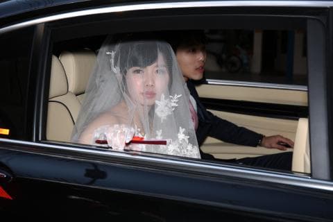 這張「婚禮」的照片是一對新人在婚禮當天，乘坐一輛黑色豪華轎車，新郎坐在副駕駛座，新娘坐在後座，新娘穿著婚紗，頭戴頭紗，新郎穿著西裝，面帶笑容。