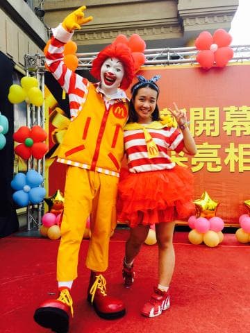 這張「麥當勞」的照片是一個穿著麥當勞制服的叔叔，他戴著假髮和紅色的鼻子，正和一位年輕女孩一起微笑。女孩穿著紅色洋裝，上面有麥當勞的標誌，看起來很開心。他們站在紅色的地板上，背景是一些紅色和黃色的裝飾。