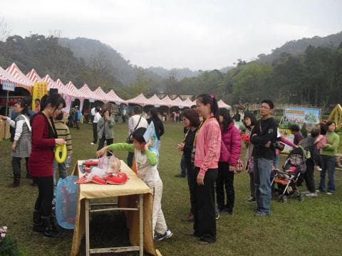 這張照片是很多人在一個露天的市場購物，有些人正在購買食物和飲料，有些人在聊天，還有一些人在看商品。照片的中心是一群人在攤位前選購商品，可以看見他們正在仔細地挑選著自己喜歡的物品。