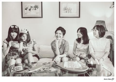 這張「四個年輕女性在客廳裡玩樂」的照片是，她們都穿著休閒服裝，其中三位坐在沙發上，另一位坐在咖啡桌旁，她們看起來很開心，並在談話或玩遊戲。