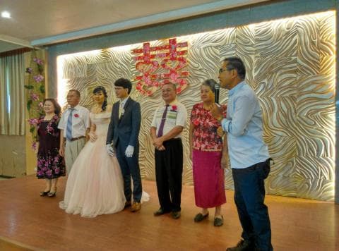 這張照片是一對新人結婚的婚禮照片，新郎穿著藍色西裝，新娘穿著白色婚紗，他們站在一起，新郎親吻著新娘。新郎的父母站在他們身後，新娘的父母則站在右側。他們身後是婚禮的背景板，上面寫著“囍”字。