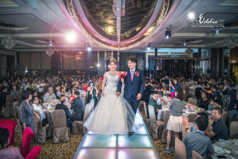 這張「婚禮」的照片是一對新人正在走入會場，他們穿著婚紗和西裝，手牽著手，臉上洋溢著幸福的笑容。賓客們坐在桌旁，熱烈鼓掌，為他們送上祝福。