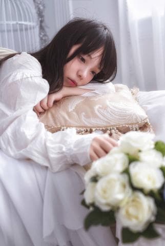 這張照片是一個年輕女子躺在床上，她穿著白色蕾絲襯衫，頭靠在一個金色花紋的抱枕上，旁邊有一束白色玫瑰花。