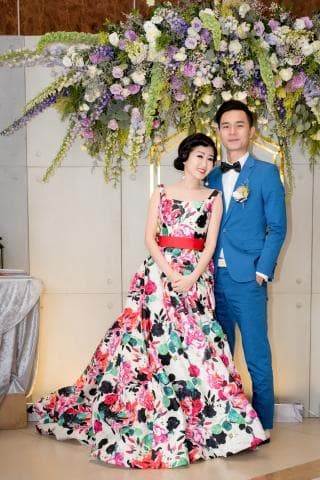 這張婚禮的照片是一個穿著花朵圖案婚紗的女子，和一個穿著藍色西裝的男子，他們站在一排花朵下面。