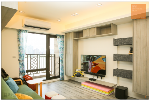 這張「客廳」的照片是一個明亮的空間，有著大大的窗戶、電視牆和舒適的沙發。牆上裝飾著木質層架，以及三層灰色的收納櫃。電視下方是彩色的坐墊，讓客廳充滿活力。