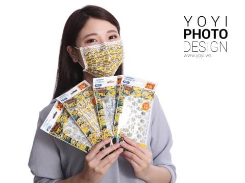 這張「戴著口罩的女生」的照片是一個女生，她戴著黃色的口罩，並且拿著幾包印有卡通人物圖案的口罩。