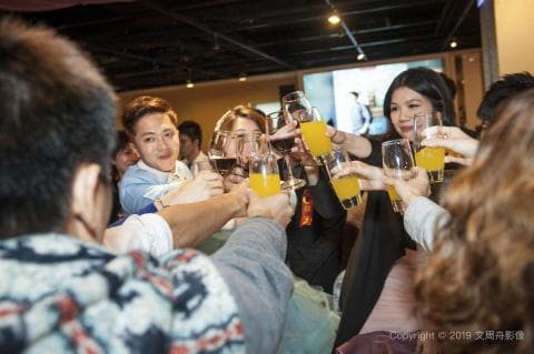 這張照片是一群朋友聚在一起舉杯慶祝的歡樂場景，照片中大部分的人都在舉杯敬酒，氣氛熱鬧又溫馨。