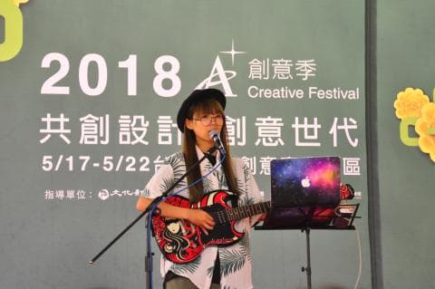 這張「2018 創意季」的照片是一個女性歌手在舞臺上演唱，她戴著帽子，穿著白色襯衫，彈著吉他，背景是綠色幕布。