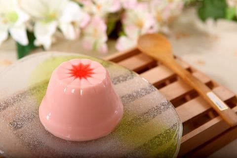 這張照片是一碗粉紅色布丁，上面有一個紅色的星星圖案。它放在一個透明的盤子上，盤子上有綠色的花紋。旁邊還有一把木湯匙。