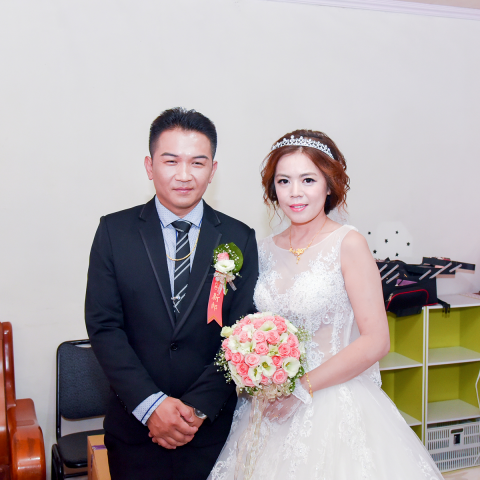 這張婚禮照片是一對新人，新郎穿著黑色西裝，新娘穿著白色婚紗，他們手上都拿著捧花，看起來很幸福。