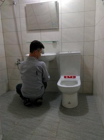 這張照片是一個浴室，照片中最重要的部分是一個穿著灰色上衣的男人，他正在修理馬桶。