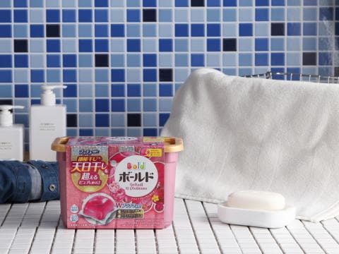 這張照片是一個放在白色瓷磚地板上的粉色洗衣球盒子，盒子上面有日文和英文的標籤。盒子旁邊有一個白色毛巾和一個白色肥皂。
