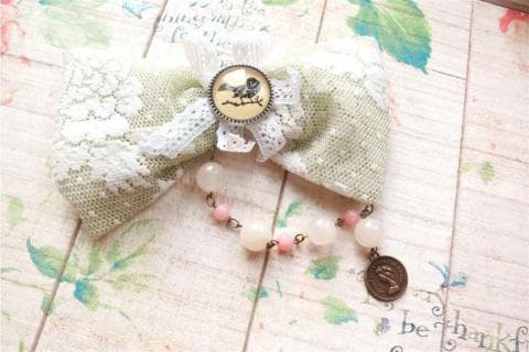 這張照片是一個綠色蕾絲蝴蝶結，上面有一個小鳥圖案的裝飾品，蝴蝶結下方連接著一串珍珠和粉紅色珠子的項鍊，項鍊的末端是一個金色的圓形裝飾。