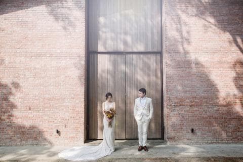 這張照片是一對新人站在磚牆前，新娘穿著白色婚紗，新郎穿著白色西裝，新娘手拿著捧花，兩個人都面向鏡頭。