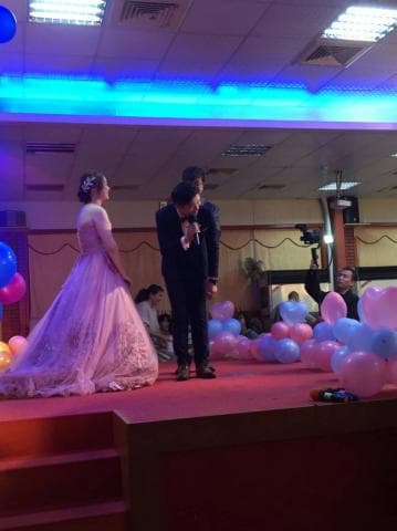 這張照片是一個婚禮現場，新郎正拿著麥克風，對著臺下的人說話，新娘穿著粉紅色的婚紗，站在新郎旁邊，看起來很幸福。