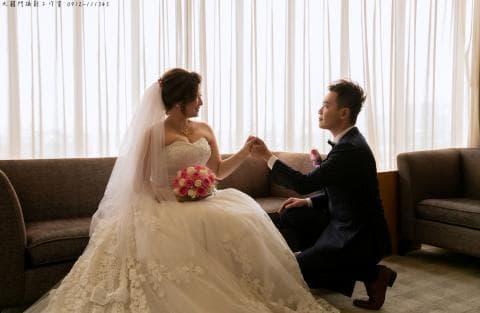 這張照片是新郎單膝跪地向新娘求婚。新娘穿著白色婚紗，手持粉色花束，臉上洋溢著幸福的笑容。新郎身穿黑色西裝，深情地看著新娘。他們之間的互動充滿甜蜜，令人感受到濃濃的愛意。