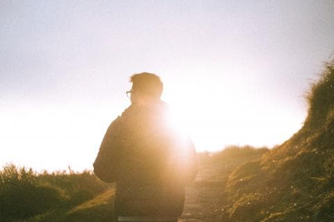 這張「一個人站在陽光下」的照片是，一個戴眼鏡的男子背對著鏡頭，站在陽光普照的草地上。