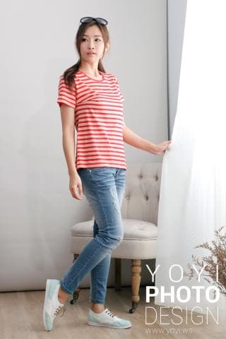 這張照片是一位年輕女性穿著紅白條紋上衣和藍色牛仔褲，她正站在房間裡，一隻腳穿著白色帆布鞋，另一隻腳則穿著白色運動鞋，並扶著一張白色椅子。