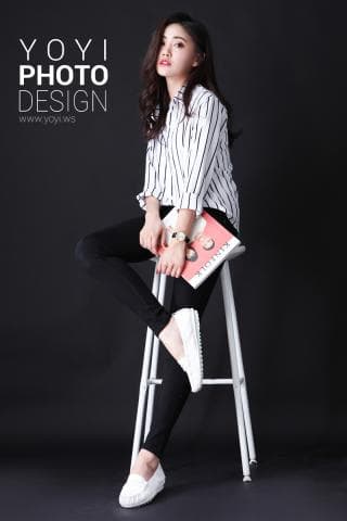 這張照片是一個女孩坐在白色凳子上，穿著白色鞋子和一條黑褲子，她拿著一本書，並且抬頭微笑著看著鏡頭。