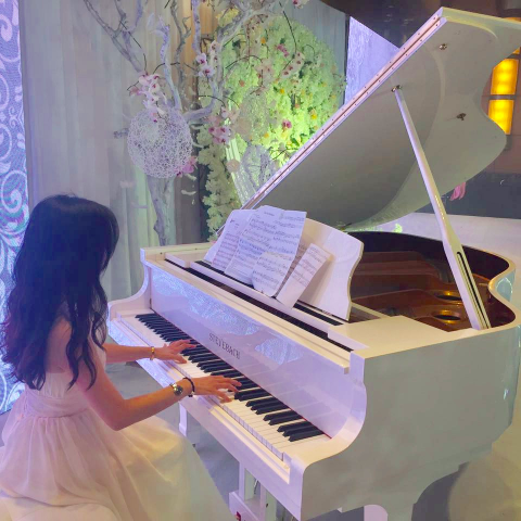 這張「一位女孩正在彈奏白色鋼琴」的照片是，女孩身穿白色連衣裙，演奏一架白色鋼琴。鋼琴旁有裝飾性的花朵和樹枝，背景是白色的牆壁。