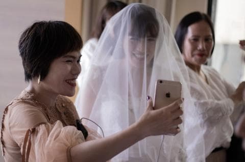 這張「婚禮準備」的照片是一個穿著婚紗的新娘，她正戴著面紗，並拿著手機自拍。旁邊有兩個伴娘，她們正在看著新娘。