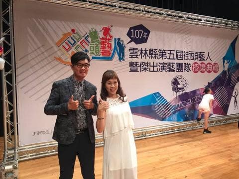 這張「舞臺照」的照片是，一位穿著西裝的男性和一位穿著白色洋裝的女性站在舞臺上，舞臺背景是一面佈滿圖案的牆。