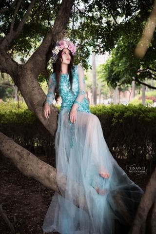 這張照片是一名女子穿著淺藍色紗裙，坐在樹枝上，背景是一片綠色的森林。