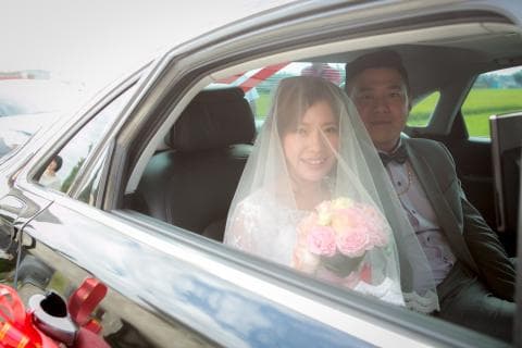 這張照片是一對新人坐在婚車裡，新娘穿著潔白的婚紗，頭紗遮住了半邊臉，笑容甜美，新郎則穿著黑色西裝，面帶微笑，兩人看起來幸福洋溢。