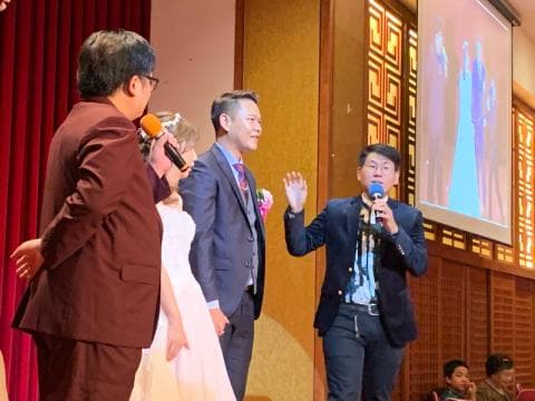 這張「婚禮」的照片是新郎新娘站在舞臺上，旁邊站著一位主持人正在講話，還有另一個賓客站在一旁，他們都穿著正式的服裝。