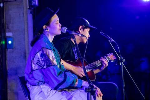 這張「音樂表演」的照片是一對男女歌手在臺上演唱，女歌手穿著亮麗的紫色外套，男歌手彈著吉他，他們都戴著耳機。