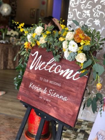 這張「婚禮歡迎牌」的照片是一塊木頭製的牌子，上面寫著「Welcome to our Wedding」，以及新人的名字「Kevin & Sienna」和婚禮日期。牌子被擺放在一個黑色的畫架上，前面擺滿了鮮花。