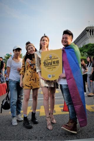 這張「四個人在街頭舉著一個標語」的照片是，一個穿著黃色洋裝的女孩子、一個戴著帽子穿著牛仔褲的女孩子、一個穿著金色連身裙的女孩子和一個穿著牛仔褲和彩虹旗的男生。