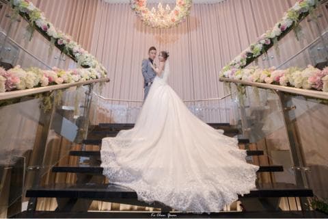 這張婚禮的照片是一對新人站在樓梯上，新娘穿著白色婚紗，新郎穿著西裝，背景是裝飾著鮮花的樓梯和牆壁。