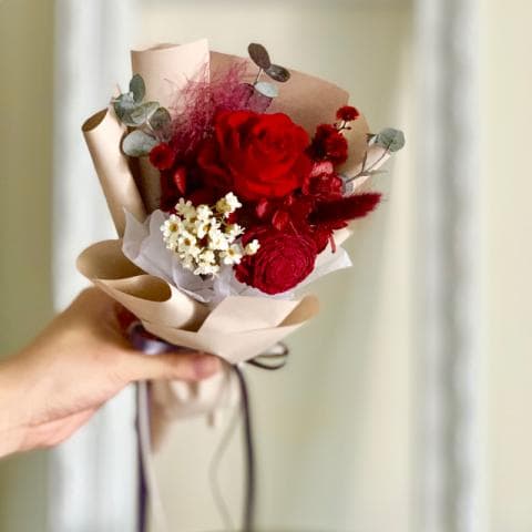 這張「乾燥花束」的照片是一束用紙包裹的紅玫瑰花束，花束中還有其他紅色的花朵和一些綠色的葉子。