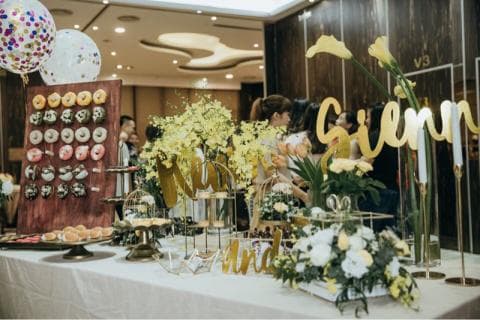 這張「婚禮」的照片是一個婚禮蛋糕桌，上面有很多甜點和花朵。桌子上有一面甜甜圈牆、一個金色蛋糕架和一個裝滿花的玻璃容器。桌上還有一對金色的「Mr & Mrs」裝飾。