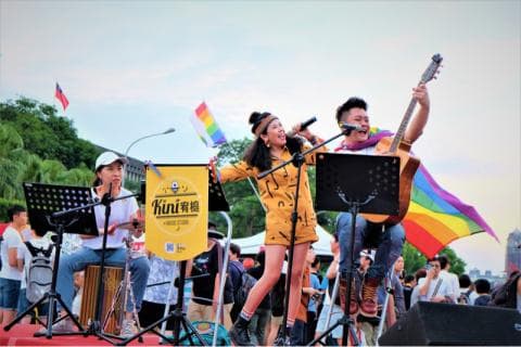 這張「街頭表演」的照片是一個樂團在戶外表演，其中有兩位女歌手，一位男吉他手，還有彩虹旗作為背景。