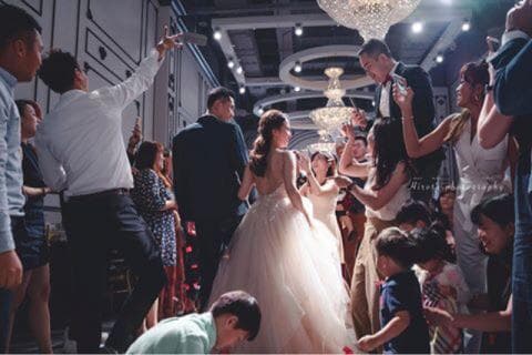 這張婚禮的照片是一對新人在眾人祝福下，準備步入禮堂的場景。 新娘穿著優雅的婚紗，新郎則穿著帥氣的西裝。 周圍的賓客們紛紛舉起手機，捕捉這難得的畫面。