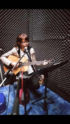 這張照片是一個年輕女子在錄音室裡唱歌和彈吉他的情景。她穿著一件棕色夾克，戴著眼鏡，正專注地看著麥克風。她手裡拿著一把木吉他，並在一個帶有三角架的立式話筒架的幫助下唱歌。