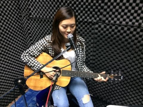 這張照片是一個年輕女子在錄音室裡彈吉他唱歌。她正在用麥克風唱歌，並專注地演奏她的木吉他。
