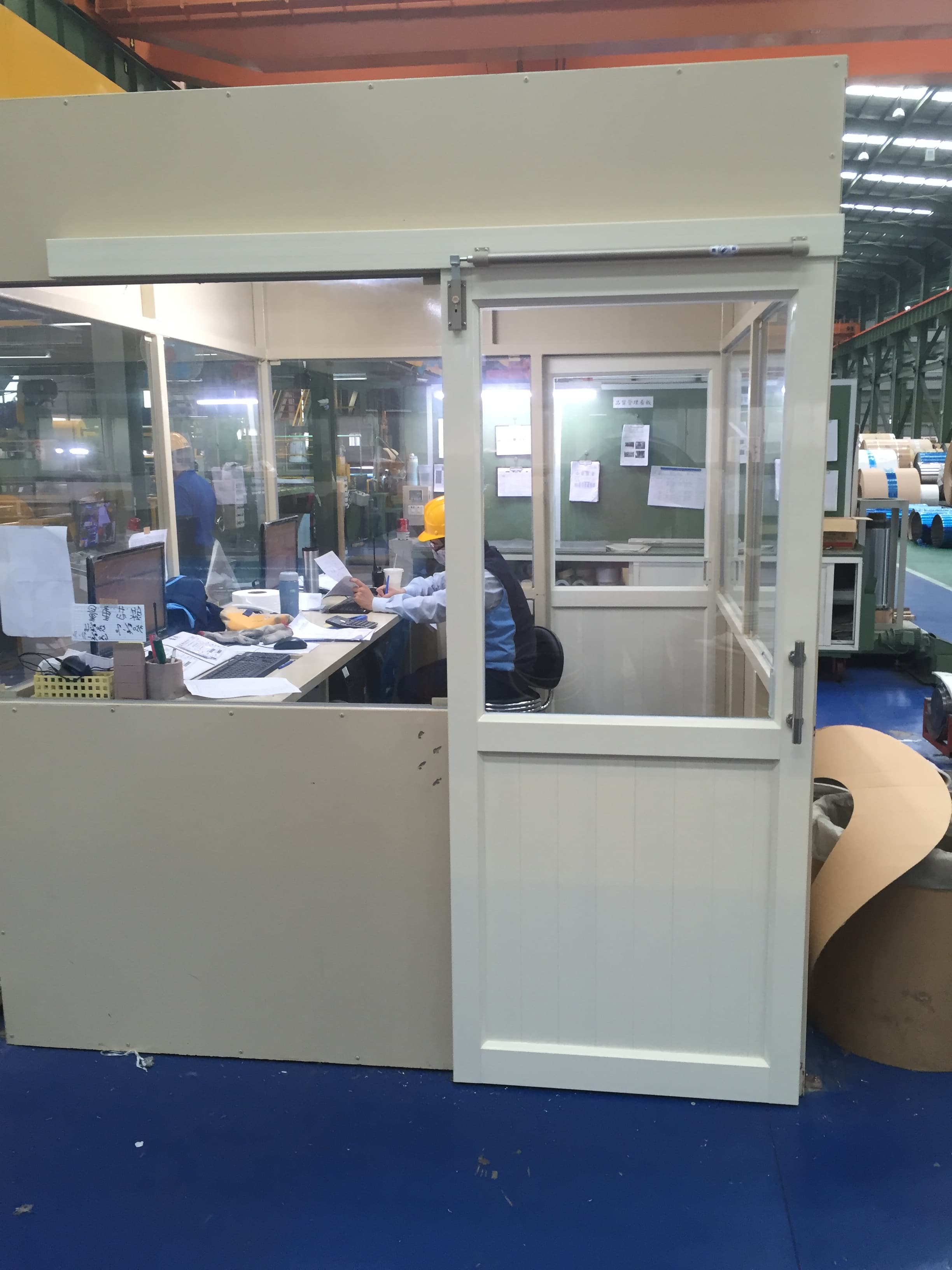 1) 照片顯示一個辦公室的內部，牆壁為淡黃色，裝設一扇白色的門，門旁有一張桌子，桌上放著電腦、文件和一個杯子